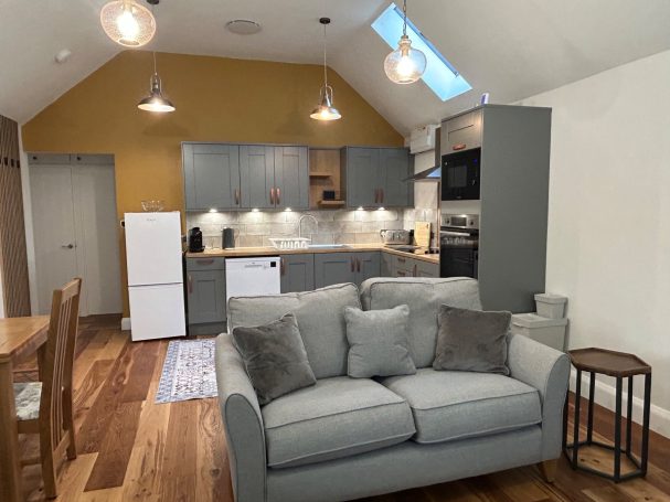 Modern kitchen and living space with a grey sofa, wooden dining table, and warm lighting.